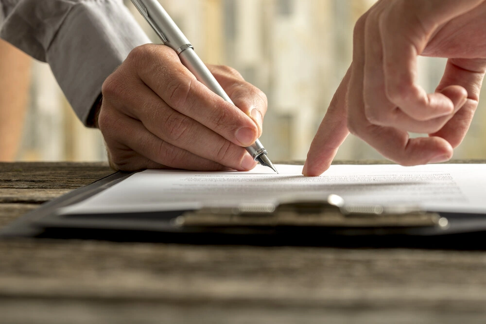 Closeup of businessman showing his new business partner where to sign an agreement or contract with fountain pen on rustic wooden desk.