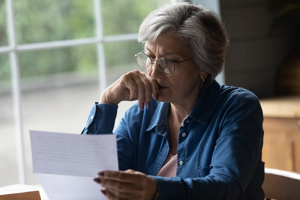 Older female sit in kitchen wear glasses reading letter received information feels upset looking concerned.